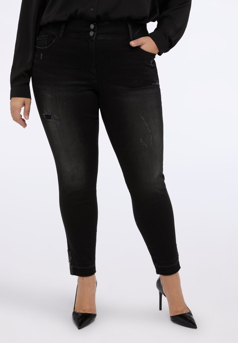 Jeans negros ajustados con un ligero desgaste, cierre de dos botones y una textura elegante. Presenta herrajes decorativos en el tobillo.