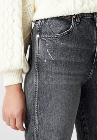 Jeans de cintura alta de color gris oscuro con una textura desgastada, que cuentan con botones de cobre, un diseño de cinco bolsillos y detalles desgastados en la tela.