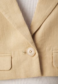Jasnobeżowa marynarka z teksturowanej tkaniny, z wyciętym klapą, pojedynczym zapięciem na guzik oraz dwoma przednimi kieszeniami. Guzik w kolorze kremowym.
