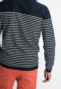 Pull bleu marine à rayures blanches, col rond et ourlet côtelé, associé à un pantalon orange. Texture détaillée visible dans le tissu.