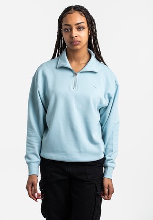 Pulóver de medio cierre en azul claro con una textura suave, corte relajado y puños acanalados. Presenta un pequeño logo en el pecho. Combinado con pantalones cargo negros.