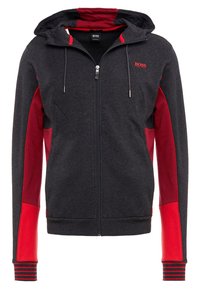Felpa grigia scura con pannelli laterali rossi, chiusura con zip, cappuccio regolabile con coulisse e polsi a righe. Tessuto morbido con una finitura liscia.