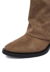 Brązowy zamszowy botek za kostkę z wydłużonym noskiem, luźnym fasonem i czarną, płaską gumową podeszwą. Gładka faktura z minimalnymi detalami.