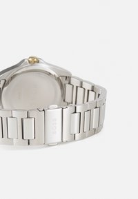 Montre en métal argenté avec un bracelet en acier inoxydable comportant des maillons polis et brossés. Accent doré sur le boîtier rond. Fond de boîtier texturé visible.