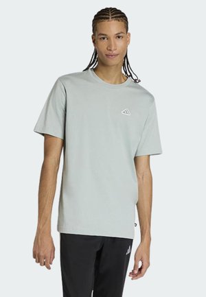 Jeune personne aux cheveux tressés portant un t-shirt gris clair à manches courtes et un pantalon noir avec un petit logo sur chaque vêtement.