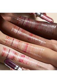 Drei Arme mit unterschiedlichen Hauttönen zeigen nummerierte Muster glänzender Lippenfarben von Nude bis Dunkelrot, mit zwei Lippenstiften, die darüber gehalten werden.