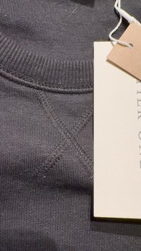 Primo piano di tessuto di felpa in cotone grigio scuro con colletto a coste testurizzato. Un'etichetta di cura è parzialmente visibile, orientata di lato.