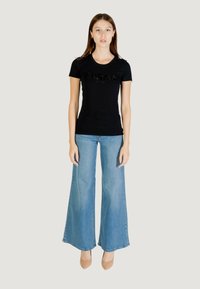 T-shirt nera a maniche corte con logo di paillettes, abbinata a jeans in denim azzurro chiaro a gamba larga. Il modello indossa scarpe con tacco color pelle, posando su uno sfondo neutro.