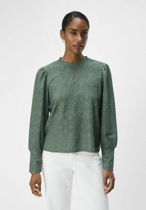 Groen kanttop met opbollende lange mouwen, ronde hals, ingewikkelde geometrische patronen en een gestructureerde afwerking, gecombineerd met witte broeken.
