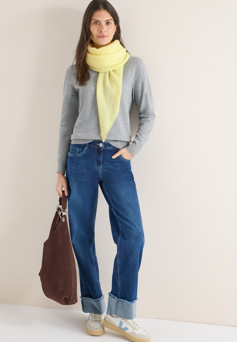 Pull gris, écharpe jaune, jean bleu avec revers en denim et baskets crème. Tenant un sac en daim marron avec un accent argenté.