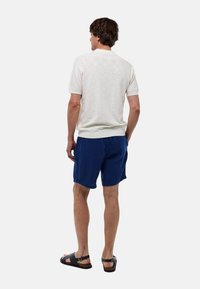 Chemise blanche texturée à manches courtes, short bleu et sandales noires. Vue de dos, mettant en valeur le col de la chemise et la taille du short.
