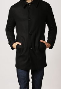 Cappotto di lana nera con colletto, bottoni frontali e tasche laterali. Presenta una vestibilità dritta e maniche lunghe, indossato sopra jeans in denim scuro.