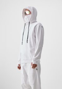 Fehér kapucnis sweatshirt, elülső kenguru zsebbel, húzózsinórral és logóval. Fehér nadrággal párosítva, amely nagy folt zsebekkel és fekete akcentussal rendelkezik.