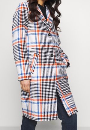 Žena nosící dlouhý károvaný kabát s oranžovým, modrým, černým a bílým pepita a kostkovaným vzorem, ruce v kapsách, doplněný tmavými kalhotami.