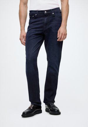 Homme portant un jean bleu foncé et des mocassins en cuir noir, debout devant un fond clair uni.