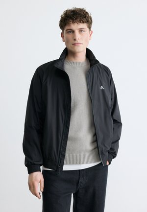 Młody mężczyzna z kręconymi włosami ubrany w czarną kurtkę na zamek, pod którą ma szary sweter i czarne spodnie, na jednolitym jasnym tle.