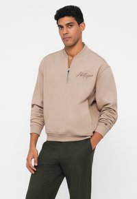 Beige mikina s krátkým zipem a žebrovanými manžetami a lemem. Na hrudi je vyšitý logo značky v písmu. Vyrobena z měkkého materiálu.