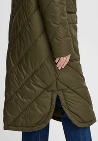 Manteau matelassé vert olive avec des coutures diagonales, un col haut et une fente sur le côté, associé à un pantalon en denim en bas.