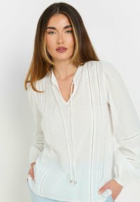 Blusa blanca ligera con escote en V, detalle de cordón, textura de puntos y acentos cortados en vertical. Mangas largas de globo con puños elásticos.