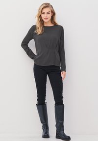 Woman with long blonde hair wearing a dark gray long-sleeve top, black pants, and black knee-high boots standing against a white wall.
