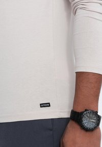 Helle beige Langarm-T-Shirt aus weichem Stoff mit einem kleinen schwarzen "Ombre"-Label am Saum. Eine schwarze Armbanduhr ist sichtbar.