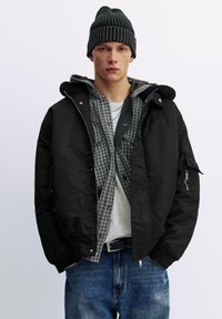 Schwarze Bomberjacke mit Kragen, darunter ein graues Karohemd, hellgraues T-Shirt, blaue Jeans und eine schwarze gerippte Mütze.