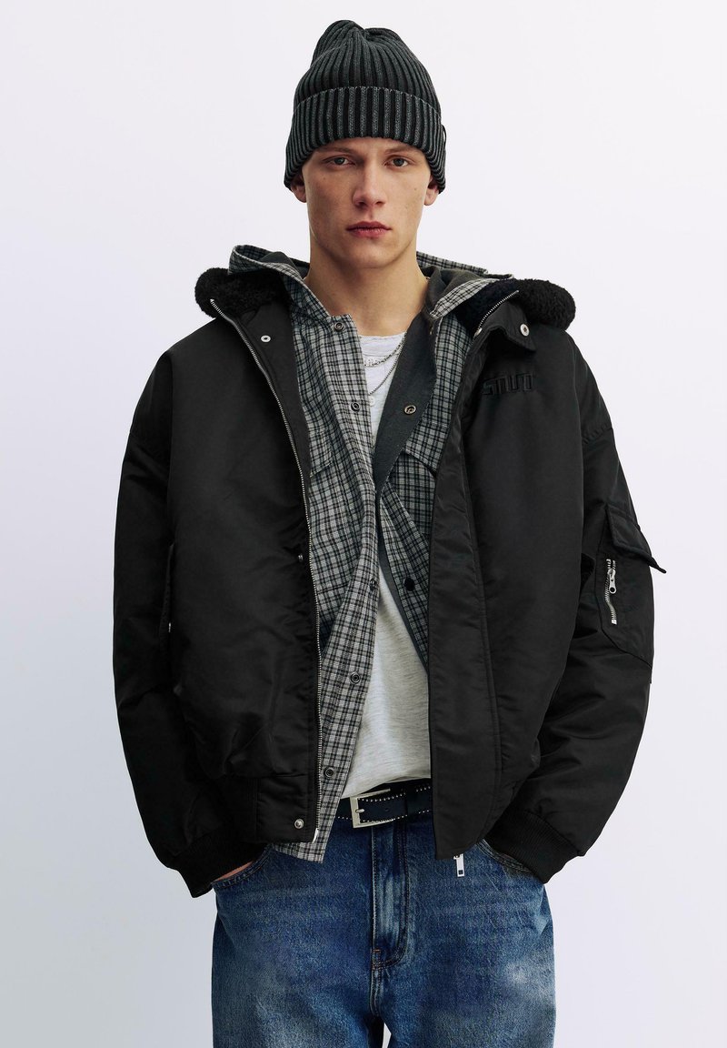 Schwarze Bomberjacke mit Kragen, darunter ein graues Karohemd, hellgraues T-Shirt, blaue Jeans und eine schwarze gerippte Mütze.