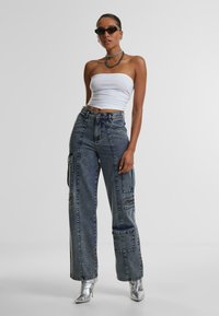 Hochtaillierte blaue Denim-Cargohosen mit Seitentaschen, kombiniert mit einem weißen trägerlosen Oberteil und silbernen Absatzstiefeln, akzentuiert durch runde Sonnenbrillen.