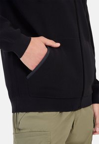 Schwarze Strickjacke mit Reißverschluss und gerippter Textur, mit seitlichem Taschen, in Kombination mit grauen Akzenten. Trägt dazu leichte grüne Cargo-Hosen.