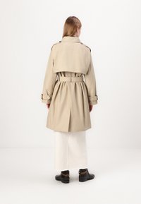 Beige trench coat s páskem v pase, ramenními epauletami a zadním rozparkem. V kombinaci s bílou sukní a černými kotníkovými botami s řetízkovým detailem.