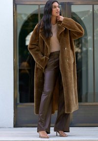 Brun faux fur frakke med dobbeltknaplukning, båret over en beige top og brune læderbukser. Klar baggrund med glasdøre.