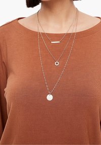 Warstwowe naszyjniki w srebrze, z okrągłym wisiorkiem, wisiorkiem w kształcie sztangi i teksturowaną dyskiem, noszone na rdzawym topie z szerokim dekoltem.