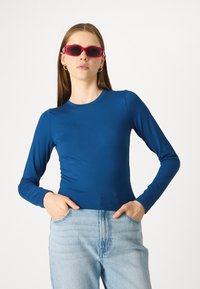 Μακρυμάνικο, εφαρμοστό μπλουζάκι από ζωντανό μπλε ύφασμα με λεία υφή, συνδυασμένο με ανοιχτόχρωμο τζιν και ροζ γυαλιά ηλίου.