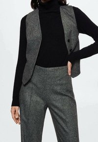 Gilet en mélange de laine gris à finition texturée, avec deux boutons noirs, associé à un col roulé noir et un pantalon gris ajusté.