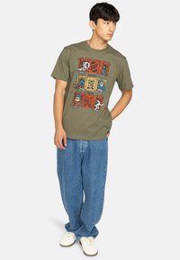 Olive green cotton t-shirt featuring a colorful graphic design with stylized characters, worn with loose blue denim jeans and white sneakers.