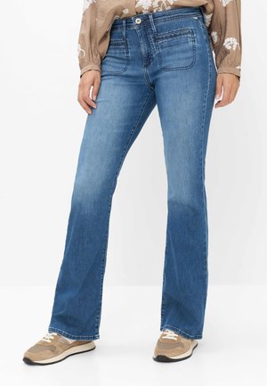 Blå denimvidde jeans med forlommelommer båret med beige mønstret bluse og beige sneakers med hvide snørebånd.