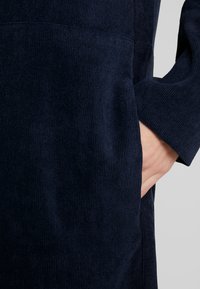 Marinblå kordflöjtstyg med ribbad textur, har en sidoficka och löst sittande ärmar. Materialet verkar mjukt och hållbart.