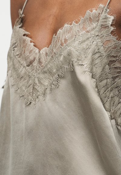 Camisolă bej din satin cu dantelă delicată de-a lungul decolteului, purtată de o persoană, cu prim-plan pe zona pieptului și a umerilor.