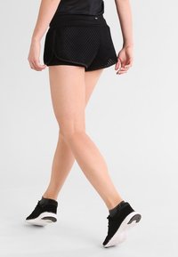 Svarta nät-träningsshorts med en bred elastisk midja och bågformad fåll, kombinerat med svarta löparskor med en vit sula.