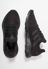 Ett par svarta stickade träningsskor med svarta sulor och tre mörka ränder på sidan, en sko sedd uppifrån och en sedd från sidan.