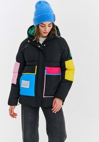 Mladá žena v čiernej prešívanej bunde s farebnými vreckami a nášivkami, modrej pletenej čiapke a tmavých džínsoch, na bielom pozadí.