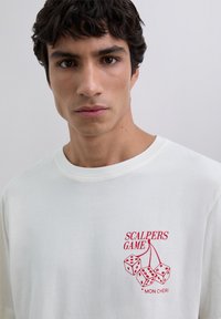 Młody mężczyzna z krótkimi ciemnymi włosami, ubrany w białą koszulę z czerwonymi kostkami do gry i napisem "Scalpers Game Mon Chéri" na piersi.