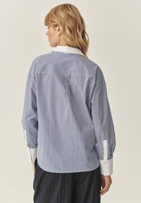 Camicia a righe blu e bianche con colletto bianco, maniche lunghe con polsini bottonati e vestibilità ampia. Presenta una spalla posteriore e righe verticali.