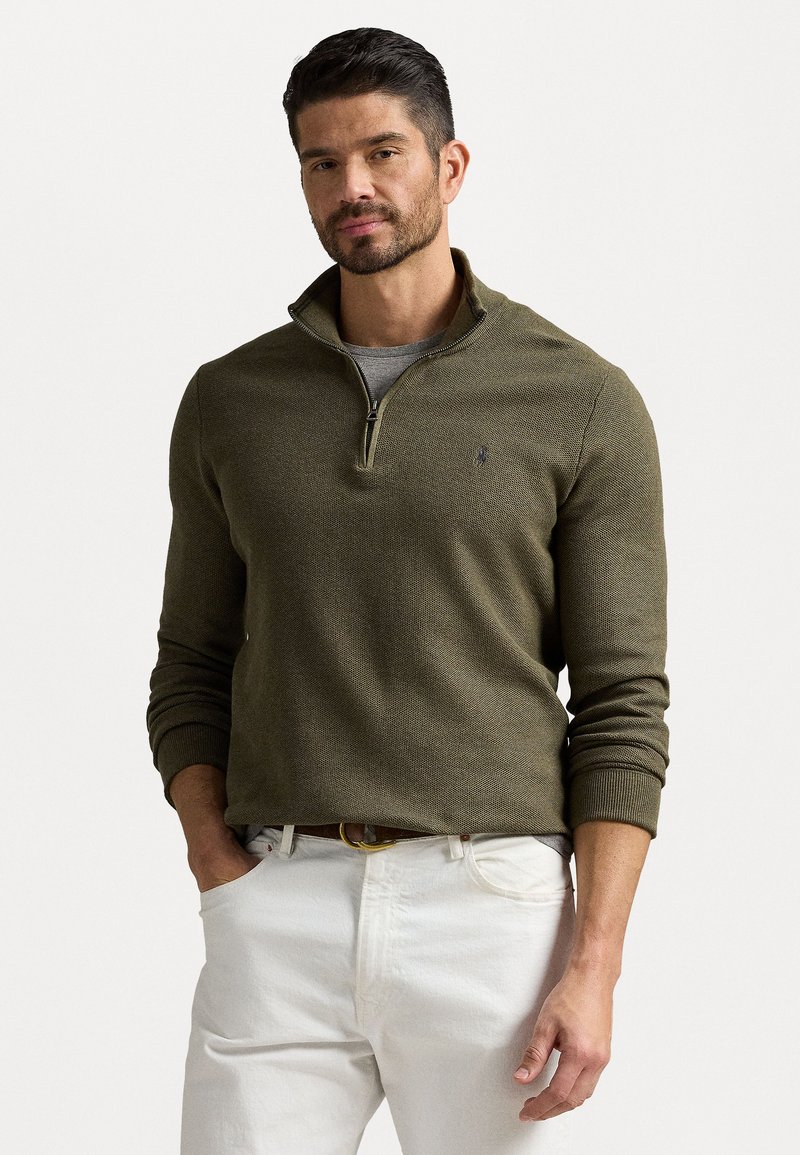 Sweater zíper verde oliva com um acabamento texturizado, apresentando um colarinho alto e punhos canelados, combinado com calças brancas.
