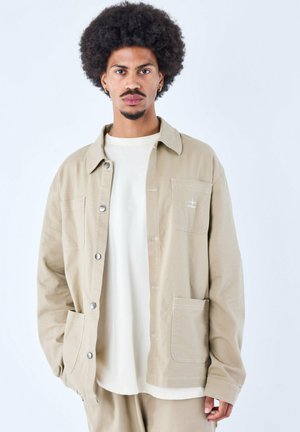 Beige katoenen jas met een kraag, voorgesloten met knopen aan de voorkant, twee opgestikte zakken en witte stiksels, gedragen over een wit T-shirt.