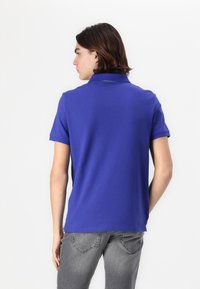 Blå polo skjorte lavet af glat stof, med krave og korte ærmer. Båret med grå jeans, vist fra bagud.