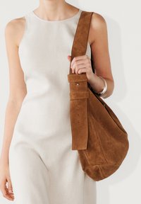 Sac d'épaule en daim marron avec une large bandoulière et un accent ceinturé. Texture douce avec une silhouette souple.