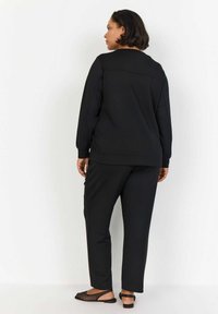 Černá mikina a kalhoty, hladký materiál, volný střih. Mezi výrazné detaily patří jednoduchý design a černé otevřené boty s perforovaným vzorem.