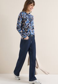 Femme marchant en souriant, portant un chemisier bleu à motifs à manches longues, un jean bleu foncé, des baskets argentées, tenant une veste beige dans une main.