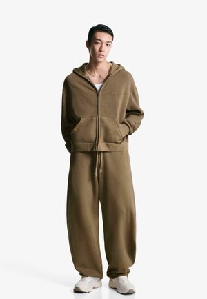 Brown zip-up hoodie with front pockets and matching loose-fitting pants. Smooth fabric, relaxed fit, paired with light-colored sneakers.